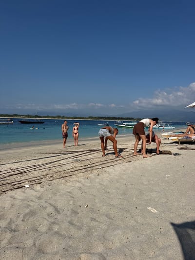 A group of people are on a sandy beach in front of a turquoise ocean under a clear blue sky. In the foreground, several individuals are engaged in various activities on the sand. One person, wearing a white tank top and brown shorts, bends over with their hands on the ground, as if picking something up or examining the sand. To their left, another person in denim shorts and a white top also bends forward, facing away from the camera. Further to the left, a man and a woman stand near the water's edge, the woman shielding her eyes from the sun with her hand. In the background, several boats are visible on the water, some docked and others further out. The ocean is calm and the water is clear. Mountains are visible in the distance on the horizon. The overall scene depicts a casual day at the beach in an tropical or subtropical location.