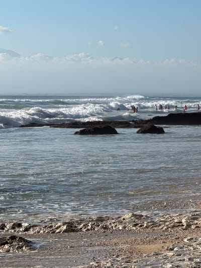 The image depicts a busy beach scene with many people swimming and playing in the ocean waves. The location is Kuta Selatan, Indonesia. In the foreground, the beach is composed of sand and rough, rocky formations partially submerged in the water. Gentle waves are lapping at the shore, with foam creating intricate patterns on the sand. Further out, larger waves are breaking, creating white surf. Numerous individuals are visible in the water, scattered amongst the waves, some appearing to be swimming or wading, while others are closer to the shore. The water is a clear blue-green, reflecting the bright, hazy sky. In the distance, a faint outline of mountains can be seen through the atmospheric haze, topped with wispy clouds. The overall scene is one of a lively and active beach environment on a sunny day.