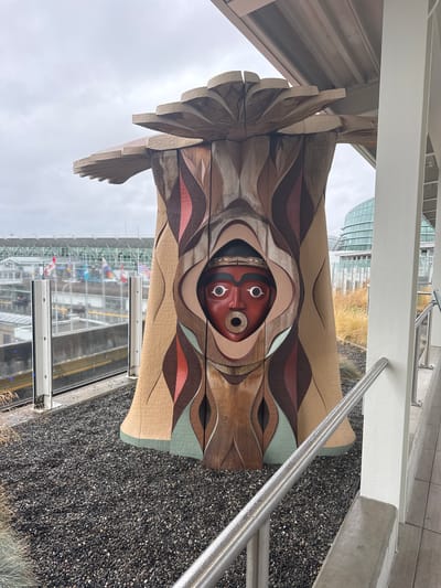 This vertical shot captures a portion of a large, carved wooden totem pole featuring a prominent, stylized face with red skin, white eyes with black pupils, and a circular opening for the mouth. The face is surrounded by intricate carvings of wavy patterns in shades of brown, beige, and red, with some accents of light green. Above the face, the totem pole exhibits feather-like carvings made of light-colored wood.

The totem pole is positioned outdoors, with a metallic handrail running diagonally across the right side of the frame. In the background, a modern building with a glass facade is visible, along with dry, golden grass. The overall atmosphere suggests an overcast day, possibly in a public outdoor space or park. The scene is oriented such that the totem pole appears to be standing upright, though the image itself is rotated. The ground beneath the totem pole is covered in dark, small pebbles. The location is identified as Richmond, Canada.