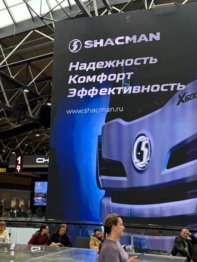 The image captures a large digital billboard displaying an advertisement for Shacman trucks in Khimki, Russia. The billboard prominently features the Shacman logo and the Russian text "Надежность Комфорт Эффективность" which translates to "Reliability Comfort Efficiency". The bottom of the billboard shows the website "www.shacman.ru" and part of a Shacman truck model, the X6000. In the background, there are architectural elements of what appears to be an indoor exhibition hall or transportation hub with a complex ceiling structure. Below the billboard, a group of people are visible, some seated and others standing. One woman in the foreground, facing left, is holding a smartphone. Another woman to her right is looking down at her phone. Further back, individuals are seated, seemingly waiting or observing. To the left of the main billboard, a smaller screen displays an advertisement for EVOLUTE, a Russian electric vehicle brand, with text in Russian. The overall scene suggests an event or a busy public space with commercial advertising.