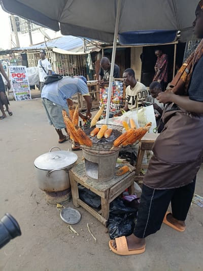 In Kabong, Nigeria, a street vendor grills corn under a large umbrella. Several ears of corn are arranged on a metal grate over a small charcoal stove. A large pot sits on another burner next to the corn. People mill around the stall, some appearing to be customers and others likely vendors. The ground is dirt and worn pavement. In the background, a collection of shops and stalls line the street, with various goods displayed. A sign with "ROLEX POS AVAILABLE HERE" is visible on the left. The scene depicts a typical urban marketplace.