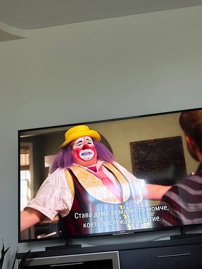 The media file shows a close-up of a television screen displaying a scene with a person dressed as a clown. The clown has purple wig, a yellow hat, and exaggerated makeup including a red nose and blue lips. They are wearing a white and yellow costume. On the television screen, there is visible text in Bulgarian: "Става дума за малкото момче, което заслужава щастие." This translates to "It's about the little boy who deserves happiness." The setting appears to be indoors, possibly a living room, with a dark framed television set. The overall tone conveyed by the clown's appearance and the text suggests a potentially emotional or narrative-driven scene, perhaps from a movie or TV show. The image is oriented sideways, with the screen taking up the right half of the frame, and the left side showing a plain, light-colored wall. A thin, dark object, possibly a stand or mount for the television, is visible on the right edge of the screen. The presence of a plant at the bottom right corner provides a hint of a domestic setting.