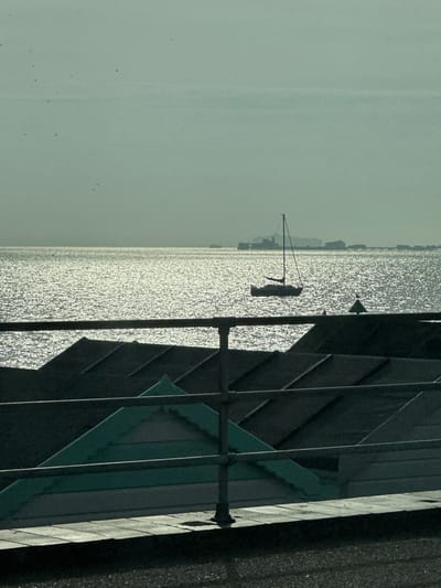 A wide-angle, backlit coastal scene captures the Thames Estuary in Southend-on-Sea, United Kingdom. In the foreground, a dark, three-bar metal railing spans the lower-mid frame above a dark ground surface. Below the railing, several beach hut roofs are visible, mostly silhouetted and dark. One prominent hut, slightly left of center, features a light teal-colored roof and a visible section of its upper wall. The midground consists of the shimmering, sun-dappled surface of the estuary, reflecting a bright, silvery light. A dark, silhouetted sailboat with a single mast floats on the water, positioned right of the frame's vertical center. Further right on the water, a dark, conical navigational buoy is present. The distant horizon features the distinct silhouette of Southend Pier extending into the estuary from the right, with various structures visible along its length. Other indistinct landforms and smaller vessels are faintly discernible along the distant waterline. The sky above is a hazy, pale greenish-grey, with several small, dark specks, likely birds, visible. No people are present.