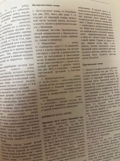 The image is a close-up, slightly angled photograph of an open book, showing two columns of Russian text. The pages appear aged and slightly yellowed. The binding of the book is visible on the left side of the image. The text is in Cyrillic script and discusses anatomical terms related to the aorta and its branches. The lighting is somewhat uneven, casting shadows from the book's spine and creases. The background is out of focus and appears to be a flat, neutral surface. There is no indication of the city of Kazan, Russia, or any forensic details.