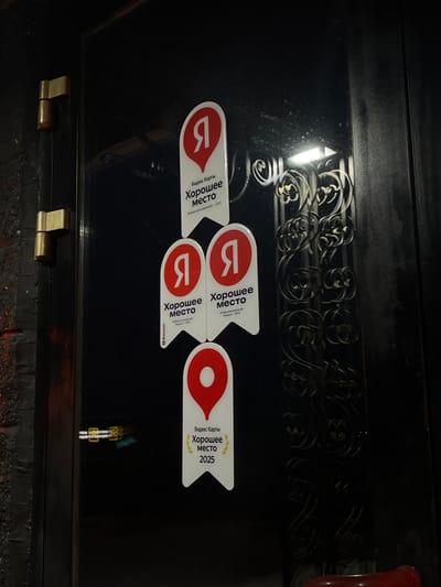 The scene depicts a dark, reflective door, likely metal, in Neftekamsk, Russia, captured in a low-light, close-up, vertical photograph. Two brass-colored cylindrical door hinges are affixed to the left frame of the door. Four white, shield-shaped award stickers from "Yandex Maps" are vertically arranged on the door's surface. The top sticker indicates "Good Place, Users' choice - 2023." Two identical middle stickers display "Good Place, Users' choice - 2022." The bottom sticker, featuring a red location pin and a laurel wreath graphic, reads "Good Place 2025," also denoting "Users' choice - 2025." All text is in Cyrillic, with prominent red Yandex 'Я' logos on the top three stickers and a red pin on the bottom. The door surface exhibits minor dust or smudges and multiple reflections: an ornate, dark, possibly metal grille or decorative element is visible on the right, a rectangular ceiling light fixture reflects from above the grille, and blurry yellow and green lights are reflected to the lower left. A dark, textured wall is partially visible to the immediate left of the door. No individuals or specific actions are present in the frame.