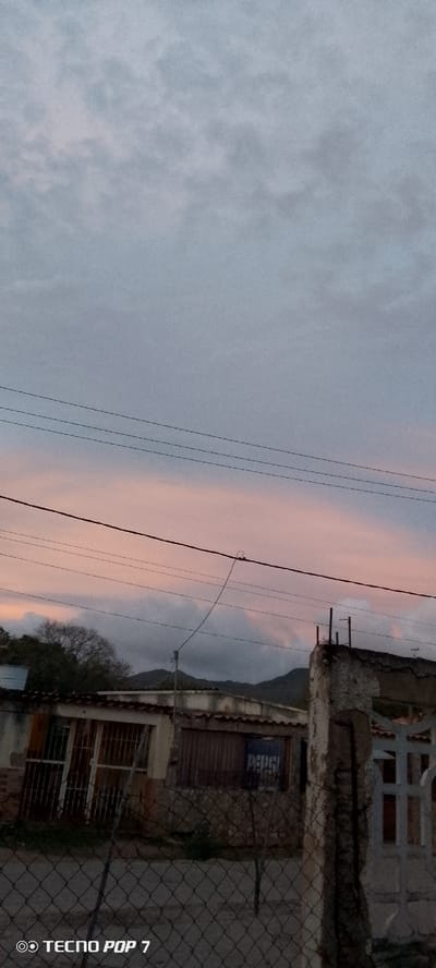 In the city of Altagracia, Venezuela, this image captures a twilight scene of a rural or semi-rural area. The sky is a muted blend of grey and soft pink, indicating the transition from day to night. Several power lines traverse the sky horizontally, supported by unseen poles. In the mid-ground, a cluster of buildings with corrugated tin roofs and what appear to be barred windows suggests a residential or utilitarian structure. Some of the buildings show signs of wear and tear. A fence constructed of wire mesh runs along the foreground, partially obscuring the view of the structures behind it. In the distance, a treeline and the outline of hills or mountains are visible against the darkening sky. A Pepsi advertisement is visible on one of the buildings. The image is watermarked with "TECNO POP 7" at the bottom left corner.