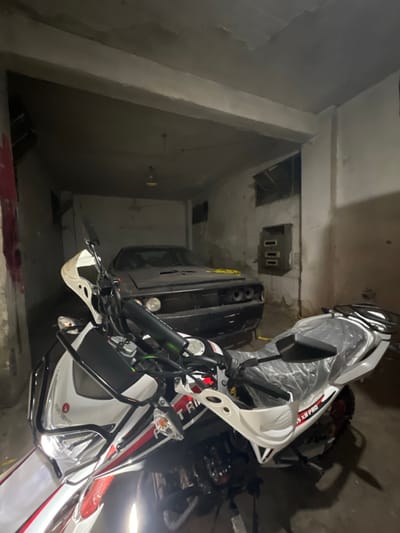 A motorbike is parked inside a concrete structure next to a black car. The motorbike is white with red and black accents. It features mirrors and a windscreen. The car is partially visible, with a portion of its rear end and side in view. The concrete structure is dimly lit and appears to be a parking garage or storage space. A small, rectangular cutout in the ceiling shows electrical connections.