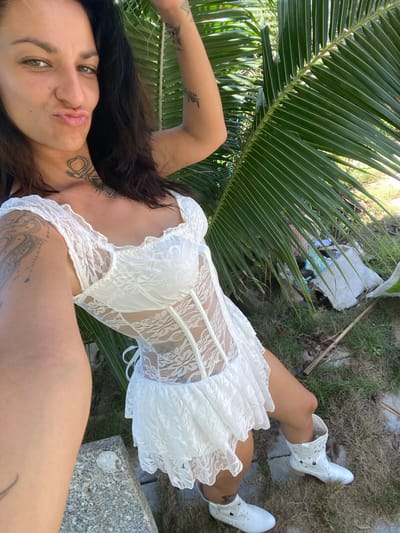 A young woman with dark hair and olive skin lies on the grass, partially obscured by a large palm frond. She is wearing a white lace mini-dress and white ankle boots. Her arms are adorned with tattoos, and she has a tattoo on her neck. The setting appears to be outdoors, with grass and some debris visible in the background. The lighting suggests it is daytime, likely sunny. The woman is looking directly at the camera with a relaxed, almost playful expression. The image is taken from a high angle, giving a close-up view of her face and upper body. The overall mood is casual and perhaps a bit bohemian.