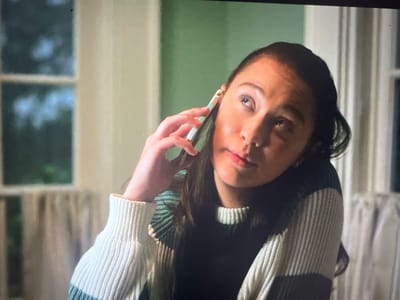 The image shows a close-up of a young woman holding a cell phone to her right ear with her right hand. She is looking upwards and to her left with her mouth slightly open. She is wearing a sweater with horizontal stripes in white, green, and dark blue. The background is a pale green wall with vertical white curtains and a window with a blurry view of trees outside. The lighting suggests an indoor setting. There is no indication of the city of Ustyvytsia, Ukraine.