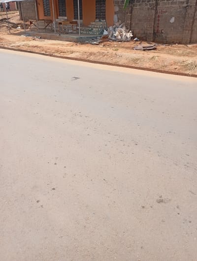 The scene depicts a static outdoor environment in the city of Akpaka Forest Reser, Nigeria. The foreground consists of a wide, light grey asphalt road showing minor surface wear and scattered dark stains. A reddish-brown dirt shoulder, approximately 1-2 meters wide, borders the road. Along this shoulder, to the right, is an accumulation of refuse, including white sacks and flattened cardboard. In the background, on the left, a building with an orange-painted facade features multiple dark, slatted windows or doors. A corrugated metal awning, supported by wooden poles, extends over a portion of this building, beneath which two light-colored wooden tables or benches are positioned. To the right, an unrendered brick wall exhibits markings in red, including numerals "+4200" and "0". No individuals or active subjects are visible. The environment appears dry and dusty under clear daylight conditions.