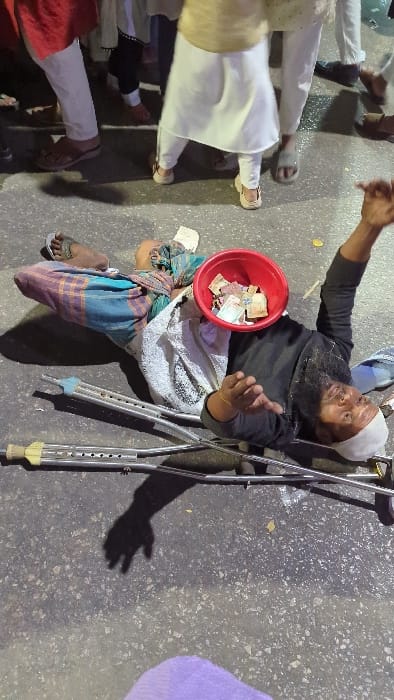 An elderly man with a beard and a white cap is lying on the ground next to a pair of crutches. He's wearing a black top and has a red bowl filled with money resting on his legs. He appears to be gesturing with his hands, possibly begging. Several pairs of legs and feet of people wearing traditional clothing are visible in the background. The scene is set on a paved surface.
