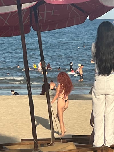 The image depicts a beach scene in Da Nang, Vietnam, with the ocean in the background and a sandy beach in the foreground. Several people are visible in the water, some swimming and others standing. A woman with long, dark hair is standing on the right side of the frame, facing away from the camera. In the middle of the frame, a woman with curly red hair is walking on the beach, wearing a black bikini. Two wooden poles, likely part of a beach umbrella or structure, are in the foreground. The water is blue with some waves. The sky is not visible.