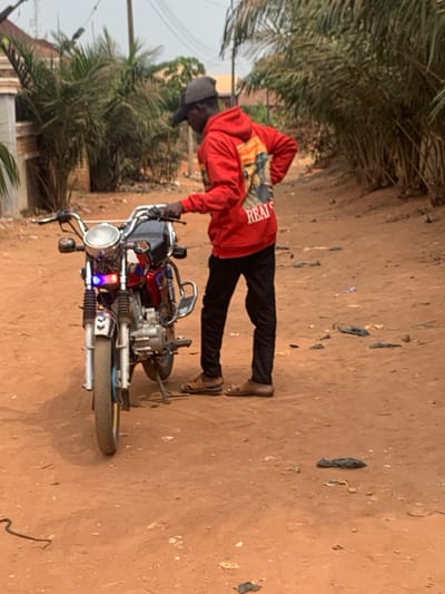 An outdoor scene in Otuocha, Nigeria, features a male individual standing beside a stationary red and silver motorcycle on an unpaved, reddish-brown dirt road. The individual is clad in a red hooded sweatshirt with a graphic on the back depicting a figure and the visible text "REALS," black trousers, brown open-toed sandals, and a dark-colored baseball cap. His left hand rests on the motorcycle's handlebars, while his right hand is placed on his hip. The motorcycle, oriented towards the viewer, has a prominent round headlight, with blue and purple accent lights positioned below it. Its kickstand is deployed. Dense green foliage, primarily palm trees, borders the dirt road, particularly on the right side of the frame. A light-colored building is partially visible on the far left. Scattered dark debris, possibly plastic, is visible on the ground. The ambient lighting suggests clear daytime conditions.