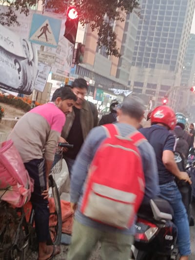 The image captures a detailed street scene in Dhaka, Bangladesh, during daylight hours, likely at a traffic intersection where vehicles are stopped. A red traffic light is visibly illuminated in the upper left, indicating a stop signal.

In the immediate foreground-left, a male individual with a beard, wearing a pink and beige long-sleeved shirt and dark pants, stands beside a black bicycle. He is observed looking downward, holding a dark, cylindrical object in his hands. A pink plastic bag, possibly a delivery carrier, is attached to the rear rack of his bicycle. Adjacent to him, a second male, dressed in a dark jacket over a dark shirt, also directs his gaze downwards, appearing to interact or observe the same point as the first individual.

In the foreground-right, a third male is seen from the back, wearing a light blue long-sleeved shirt, olive-colored pants, and a prominent red backpack with white accents. He is standing in close proximity to a stationary black scooter, identified by a visible "Mahindra" logo on its rear side. Directly behind this individual, a fourth male is seated on the black scooter, wearing a dark blue short-sleeved shirt, blue jeans, and a red motorcycle helmet. His right hand is positioned on the handlebar.

Further in the mid-ground and background, additional male individuals on motorcycles are visible, queued in traffic. One notable rider wears a green helmet.

Above the scene on the left, a blue square traffic sign with a white border and a black pedestrian symbol is affixed to a pole. This pole is also covered with multiple white rectangular posters featuring Bengali script and portrait photographs. A low concrete median strip, adorned with orange flowering plants, runs along the left side of the street. In the background, several modern, multi-story buildings with grid-patterned facades dominate the urban skyline.