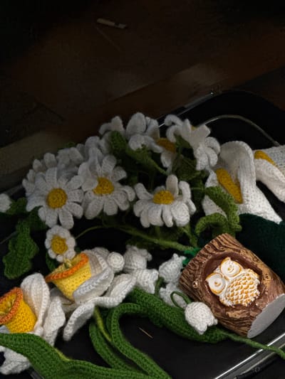 The image depicts a close-up of crocheted flowers and a decorative owl figurine arranged on a dark, flat surface that appears to be the lid of a suitcase. The crocheted items include white daisy-like flowers with yellow centers, white lily-of-the-valley flowers, and green stems and leaves. There are also crocheted daffodils with yellow and orange cups. The owl figurine is small, ceramic, and features an owl nestled in a tree-like cavity. The background is dark and out of focus, showing what appears to be a wooden surface or wall. The scene is indoors. The image does not provide any information about the city of Tbilisi, Georgia, or any specific location within it.