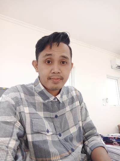 A man poses indoors wearing a blue and white plaid shirt. His hair is dark and styled upward. The background features a white wall with a window and an air conditioning unit. A stack of papers is visible in the lower right corner. The man has a slight mustache and beard. He looks directly at the camera with a neutral expression.