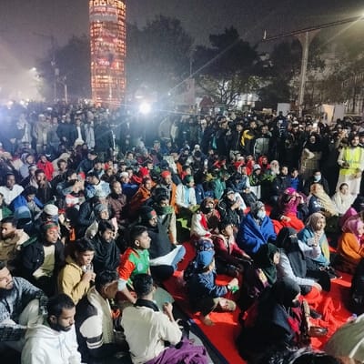 A large crowd of people is gathered outdoors at night. Many are sitting on blankets or the ground, while others are standing behind them. A tall, illuminated structure with vertical text in a foreign language is visible in the background. The people appear to be observing something. A person wearing a reflective vest stands at the edge of the crowd. The scene suggests a vigil, protest, or an outdoor event.