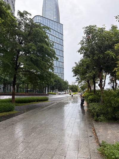 The image depicts a wet, tiled pedestrian walkway in a city park. Tall, modern buildings with glass facades loom in the background, suggesting an urban environment. Green trees and manicured bushes line the walkway, with lampposts visible interspersed throughout the park area. The walkway is wet, reflecting the overcast sky and indicating recent rain. Several people are present, including a person walking with an umbrella, a person on a bicycle, and individuals on motorcycles. Parked cars are visible on a street bordering the park. The scene conveys a sense of a peaceful, albeit damp, day in a well-maintained city park with modern architecture.