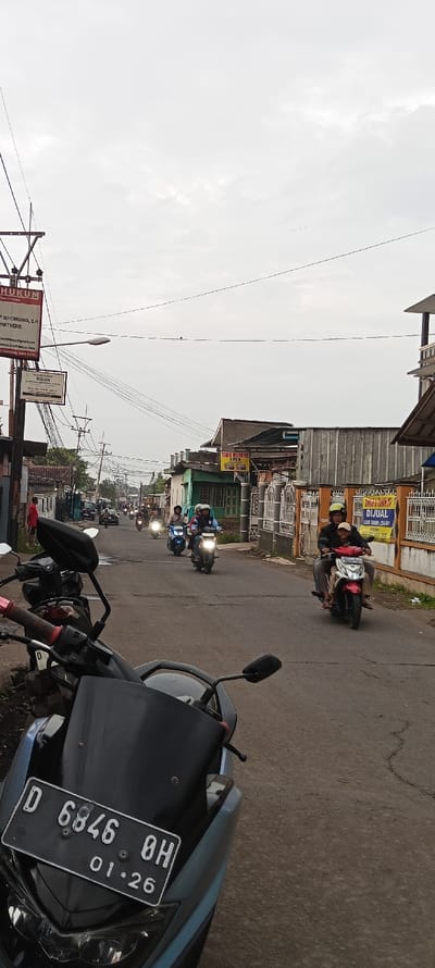 The image shows a street in Cangkuang, Indonesia (latitude: -7.054, longitude: 107.554). There are several motorcycles parked on the side of the street, and a few more are traveling down the road. The street is lined with buildings on both sides, and the sky is overcast. The street is paved with gray asphalt and has visible cracks in the asphalt. A black motorcycle with a blue front fender is parked in the foreground. The license plate reads "D 6846 8H 01.26." Three people are riding motorcycles, and one is standing on the sidewalk. Two of the motorcycles are blue, and one is red. There are several signs in the scene, including a "TAHU GORENG" sign, and a "DIJUAL" sign. The image was taken in the late afternoon, as the sun is setting.