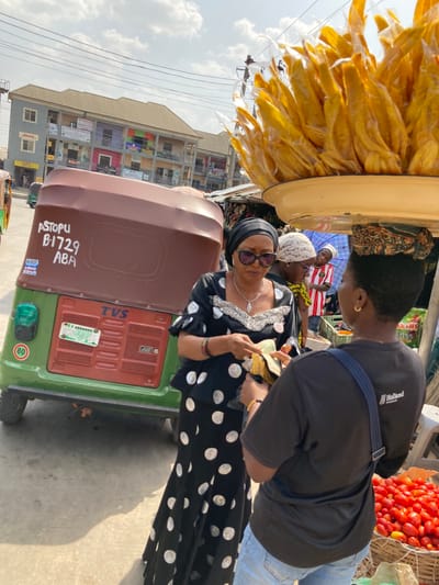 The image depicts a daytime street market scene in Aba, Nigeria. In the foreground, an adult female wearing a black top and long skirt with white polka dots, a black headwrap, and sunglasses is holding currency notes, engaging in a transaction with another individual. This second individual, facing away from the camera, wears a black t-shirt with "Holland" text and light blue jeans, and carries a large, light-yellow tray laden with individually plastic-wrapped yellow items (likely snacks) balanced on their head. A basket filled with red tomatoes is visible near their feet.
Immediately behind the polka-dotted woman, a green and brown motorized tricycle (keke napep) is parked. It bears the markings "ASTOPU B1729 ABA" in white paint on its rear, indicating its operational location. The tricycle is a "TVS" brand and displays a green Nigerian license plate reading "MSD ANAMBRA," along with a circular "80" speed limit sign. In the background, a multi-story building with balconies and various commercial signs, including one for "DHL," lines the street. Other individuals are visible in the mid-ground and background, contributing to the market atmosphere. Overhead power lines are visible against a clear, bright sky.