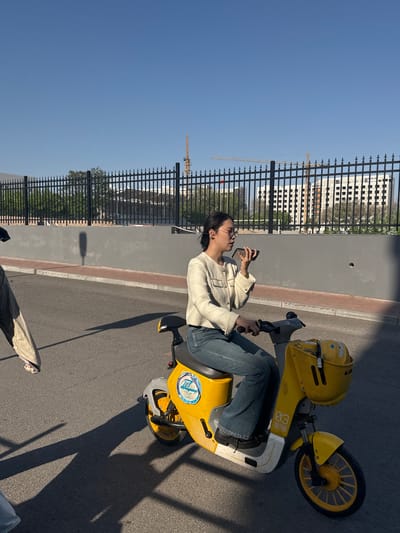 A young woman sits on a yellow electric scooter on a paved surface. She wears jeans, a light-colored tweed jacket, and glasses. She holds a cell phone in her left hand and the scooter's handlebars in her right. A black metal fence lines the left side of the frame, behind which is a multi-story building with a construction crane visible in the background. The sky is clear and blue. The scooter casts a shadow on the paved surface. Given the visible identifiers, it is highly possible that this photograph was taken in or near Zhongtun, China.