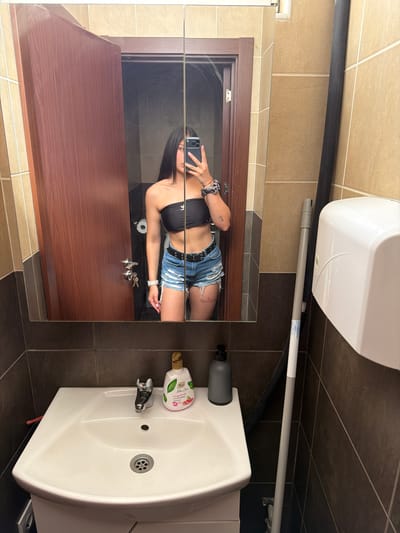 The image depicts a reflection in a bathroom mirror, showing a young woman with long dark hair wearing a black tube top and denim shorts. She is holding a smartphone and taking a selfie. The bathroom features a wooden door, a sink with a faucet, a bottle of liquid soap, and a dispenser for paper towels. The walls are tiled in beige and dark brown. A mop handle is visible leaning against the wall. The scene is set in a bathroom, but there is no information in the image that would allow for the determination of a specific city or country, including Gabrovo, Bulgaria.