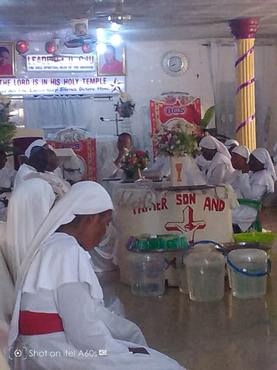 A low-angle indoor shot depicts a religious gathering, likely in a church, somewhere in Lagos, Nigeria. A group of people, mostly adults, are clad in white robes and head coverings. The backdrop features a sign that reads "THE LORD IS IN HIS HOLY TEMPLE" and another with portraits and the words "LEADER O O ERU THE SULA SPIRITUAL HEAD OF THE UNIVERSE." The altar is decorated with flowers. Clear plastic containers are on the floor. A sign reads "FATHER SON AND" with a red cross design. A gold-painted pillar with a purple spiral pattern is visible. A clock is on the wall.