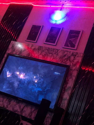 The image depicts a dimly lit room, possibly a personal entertainment space, featuring a television screen displaying a scene of destruction and fire, possibly from a movie or game. Above the television, three framed black and white photographs are displayed on the wall. The room is illuminated by a strip of pink LED lights running along the top of the wall and a blue-toned spotlight. The walls have a textured finish, and the furniture or decorative elements visible are in dark tones with marble-like patterns. The overall ambiance is dramatic and atmospheric, enhanced by the interplay of colored lights and the content on the screen. The location in New Stadium, Nigeria is not discernible from the visual information provided.
