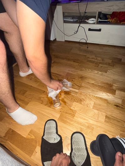 The image depicts a domestic indoor scene, likely a living room or bedroom with a wooden floor. The focus is on the lower legs and feet of at least two individuals. One person, wearing dark blue shorts and white socks, is actively cleaning up a spill from a glass on the floor. The glass appears to be partially overturned, with liquid and possibly ice cubes scattered around it. This individual is using a white paper towel to soak up the spill.
In the foreground, two pairs of footwear are visible on the floor: a pair of black slippers and a pair of black Adidas slides. The presence of these items suggests a relaxed, at-home environment. In the background, a wooden furniture unit, possibly a TV stand or cabinet, is visible. Cables are seen hanging from the unit, indicating the presence of electronic devices. The overall impression is of a casual setting where an accident has occurred and is being addressed. There is no discernible information within the image to suggest a location in Podgorica, Montenegro.
