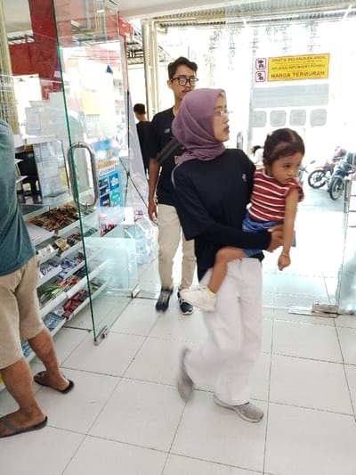 A woman wearing a black shirt, white pants, and a purple hijab, is holding a young girl wearing a red and white striped shirt, and blue jeans. The woman is walking towards the camera with the child in her arms, and both are looking at the camera. Behind them is a man wearing a gray shirt, black pants, and glasses. The man is walking out of the store, and is also looking at the camera. There is a store shelf in the background, stocked with various items.  The environment is a shop or store, with a glass door on the right side of the image.  There is a sign on the glass door that says "GRATIS BELANJAAN ANDA DI WAJAH INFORMASI PONSEL HARGA TERMURAH" (Free Your Shopping on Our Mobile Information Center, Cheap Prices). The location is -6.753 latitude and 111.041 longitude in Pati, Indonesia.