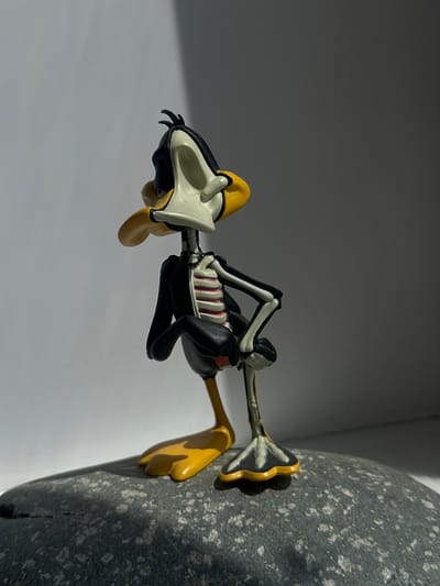 This is a full-body, eye-level, close-up, studio shot of a Daffy Duck figurine posed on a dark, textured rock. The figurine is a stylized representation of the character with a skeletal structure exposed. Daffy Duck's black feathers are visible on his head and arms, with a white skull-like face and a yellow beak. His ribcage is exposed, showing pink ribs and a dark torso. His arms are bent, and his hands are clasped behind his back. His legs are yellow, with visible bones, and his feet are webbed and yellow. The background is a gradient from light gray to dark gray, with a distinct shadow cast from the left side. The lighting appears to be directional, creating highlights and shadows on the figurine and the rock. There is no discernible connection to Khabarovsk, Russia.