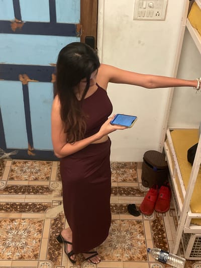 In Vagator, India, a young woman with long, dark hair stands in what appears to be a dormitory or hostel room. She is wearing a maroon two-piece outfit, with a halter-neck crop top and a long, flowing skirt. She is looking down at a smartphone held in her right hand, with her left arm extended. Her left wrist is adorned with bracelets. To her right, a bunk bed with yellow bedding is visible. On the floor, near the bunk bed, are a pair of red slip-on shoes, a black flip-flop, and a water bottle. The room has tiled flooring with an ornate pattern and a blue door is visible on the left side of the image. A light switch is mounted on the wall.