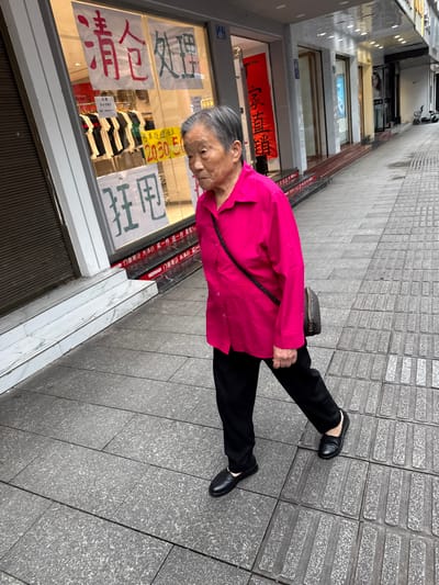 An elderly woman wearing a bright pink jacket, black pants, and black shoes stands on a paved sidewalk in what appears to be Jiangjin, China. She is positioned in front of a store with signs written in Chinese characters displayed in the window, one specifically marked "清仓处理" (clearance sale) and other phrases. She has a black purse slung across her body. The environment suggests a commercial street with shops lining the sidewalks, and the pavement indicates an urban setting.