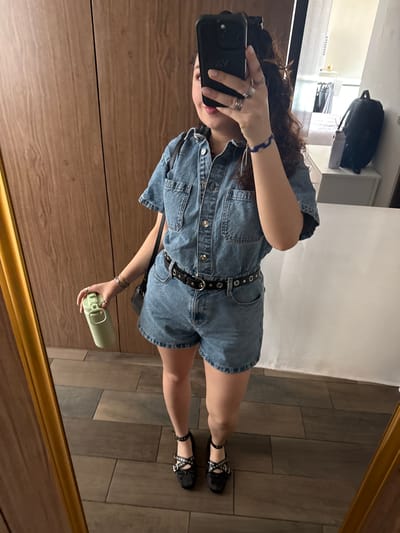 A person is seen in a mirror selfie, holding a cell phone in their left hand. The person is wearing a denim jumpsuit with a black belt. They are also wearing black studded sandals. In their right hand, they hold a green water bottle. The reflection shows a wooden floor. Other objects in the background of the reflection include furniture and a backpack. It cannot be determined that the location is in Mexico City, Mexico.
