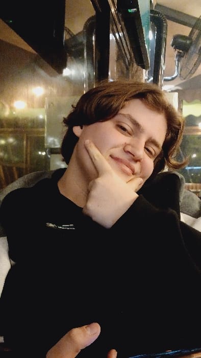 A person with short brown hair and a black hooded sweatshirt smiles and poses for the camera. Their left cheek is playfully pinched by a pointing finger. The image is taken from a low angle, with a glossy table and a fan visible in the background. The lighting is warm and indoor. The sweatshirt's sleeve and someone else's hand are partially visible on the right side of the frame.