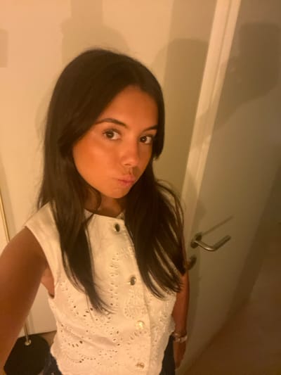 An indoor, waist-up self-portrait depicts a young adult female, standing with her upper body angled slightly to the right, looking directly at the camera. She has long, dark hair, a tan complexion, and brown eyes. Her lips are puckered in a "kiss" or "duck face" expression. She wears a white sleeveless blouse with an eyelet embroidery pattern and a vertical row of gold-toned buttons down the front. A silver-colored watch is visible on her left wrist. The background features a plain, light-colored wall to the left and a light-colored door with a horizontal silver-toned handle to the right. A dark, thin, vertical object is partially visible at the bottom left. The environment is illuminated by warm-toned artificial light, casting subtle shadows. The image lacks specific identifying features to determine a precise location within Lordelo do Ouro e Massarelos, Portugal, beyond being an interior space.