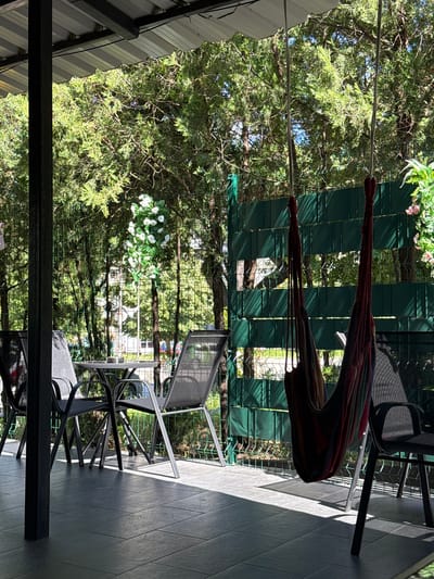 An outdoor patio area is visible, featuring dark gray tiled flooring and a dark gray metal roof supported by a black pillar on the left. A round metal table with four dark gray metal chairs is set up, with an ashtray on the table. A multi-colored hammock, made of fabric with red, green, and brown stripes, hangs from a metal stand to the right of the table and chairs. Beyond the patio, a green fence encloses a grassy area with trees and foliage. A cluster of white artificial flowers hangs from a tree on the left side of the fence. In the background, through the fence and trees, a street with passing cars and buildings can be seen, suggesting an urban environment. The scene appears to be a seating area outside a residential or commercial establishment. The image does not contain any people. The location is identified as being in Shumen, Bulgaria.
