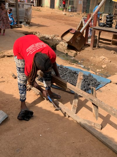 This image depicts a street scene in Sabon Tasha, Nigeria, likely during daylight hours given the strong shadows. The central subject is a person wearing a red t-shirt with white text and patterned trousers, actively engaged in some form of work. They are holding a hammer with a blue handle, appearing to be striking something out of frame. Their hair is braided and cascades down their back. Beside them, a pile of dark, granular material, possibly charcoal or coal, is spread out on a blue tarp, supported by wooden planks.
In the background, there are hints of a marketplace or a busy street. A metal structure, possibly part of a stall or cart, is visible. Further in the background, other individuals can be seen, including two young girls, one wearing a pink dress and the other a blue and patterned fabric. The ground is a dusty, reddish-brown earth, typical of many areas in Nigeria. The overall impression is one of everyday life and commerce in a bustling Nigerian community.
Visible text on the person's red t-shirt includes "VISIT SALAMA CENTER," a name, and what appear to be phone numbers and a location indicating "Kakuri, Kaduna." The presence of this text suggests the person may be associated with or promoting the Salama Center. The scene seems to be outdoors, with an arid or dry climate suggested by the dusty ground.