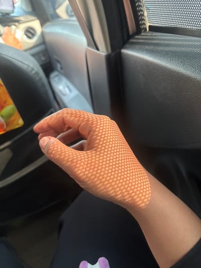 The image shows a close-up of a person's hand and forearm resting on what appears to be a dark-colored seat inside a vehicle. The hand is illuminated by sunlight filtering through a patterned mesh screen, casting a distinct hexagonal shadow pattern across the skin. The background is blurred, but hints of car interior elements like a window, door panel, and possibly a dashboard are visible. The scene is not specifically located in Tangerang, Indonesia, as there are no visible geographical markers to indicate this.