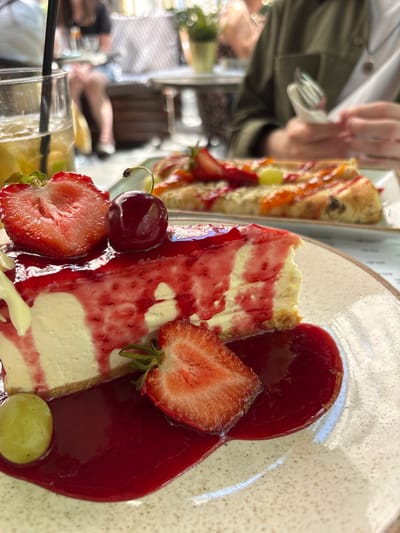 An outdoor dining scene in Belgrade, Serbia, features a close-up of a slice of cheesecake on a speckled plate. The cheesecake is topped with red berry sauce and garnished with a halved strawberry, a whole cherry, and a green grape. In the background, a person is visible holding a fork and napkin, and another person is seated at a table with a drink. The setting appears to be a restaurant or cafe with blurred foliage and outdoor seating.