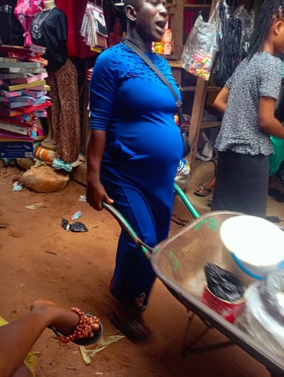 In Nkpor, Nigeria, a woman wearing a blue dress is pushing a wheelbarrow. She is in a market, with stalls and goods visible around her. The wheelbarrow contains various items, including a white bucket and a red container with black material. Another woman, wearing a patterned top and a black skirt, stands nearby. The ground is sandy and there are some discarded plastic bags.
