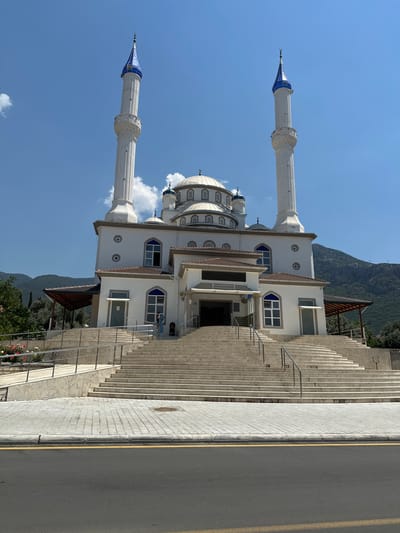 The image shows a mosque with two minarets, a dome, and a white facade, set against a backdrop of lush green mountains under a clear blue sky. The mosque features blue-tipped minarets and intricate architectural details on its dome. A wide, long staircase made of stone leads up to the entrance of the mosque. A paved walkway with a yellow line borders the staircase and a road.
One person is visible near the entrance of the mosque, suggesting a calm and ordinary day. The bright sunlight and clear sky indicate it is daytime, likely midday or early afternoon. The presence of a mosque and the mountainous landscape are characteristic of locations like Fethiye, Turkey. There is no visible text in the image.