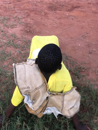 An overhead shot captures a person lying or sitting on the reddish-brown soil in Ukwu-Abwa, Nigeria. The individual, likely a child or young person, is viewed from directly above, with their head positioned towards the upper left of the frame. They are wearing a bright yellow long-sleeved shirt. Over the yellow shirt, a light-colored, possibly beige or tan, distressed garment with prominent stitching and frayed edges covers their lower torso and legs, resembling worn shorts or a skirt.
The person's head, covered in short, tightly curled black hair, is visible. Their left ear is partially seen. A dark-skinned arm extends outwards towards the right, resting on patches of green grass interspersed with the soil. The ground is a mix of bare reddish-brown earth and sparse green foliage, characteristic of an outdoor, possibly rural, setting. Small stones and dry plant matter are scattered across the soil. The lighting suggests it is daytime, though the exact time is not discernible. No specific activity beyond resting or lying on the ground can be clearly identified, and emotions are not visible. There is a small white rectangular patch on the yellow shirt, but no legible text is visible on it or elsewhere in the image.