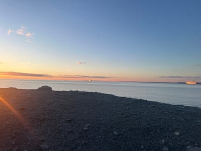 A wide shot captures a tranquil seascape at sunset, with a rocky, dark grey shoreline dominating the foreground. The sun, low on the horizon to the left, casts a warm, orange glow across the water and the sky, creating a soft gradient of colors from deep orange near the horizon to a pale blue at the top. Wispy clouds are scattered across the sky, tinged with pink and orange from the setting sun. Several vessels are visible on the horizon. To the far left, a small sailboat is faintly visible. Further to the right, a larger, yellow-hued vessel appears to be a ferry or cargo ship. On the far right side of the image, a brightly lit passenger ship, possibly a cruise liner, sails across the water. The ocean is calm, reflecting the colors of the sky. The rocky shoreline is composed of small to medium-sized stones, with a few larger boulders present. The scene appears to be a coastal area, likely an outcropping or cliff edge. There are no people visible in the image. The overall impression is one of serene natural beauty at the end of the day.