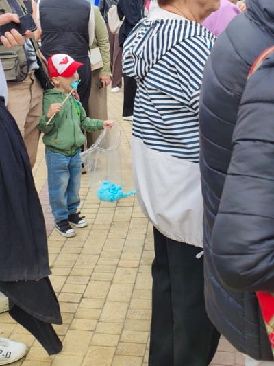 In Kirovsk, Russia, a young boy wearing a green hoodie and blue jeans stands on a paved area, holding a stick with a blue cotton candy and a plastic bag containing more blue cotton candy. He is also wearing a red and white baseball cap. People are gathered around him, with some individuals partially visible in the frame. The environment appears to be an outdoor public space, possibly a street or a park, with a tiled pavement.