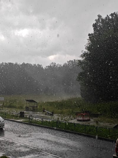 The image depicts a scene of heavy rainfall in a residential area, likely a playground or park, in Chelyabinsk, Russia. White specks, representing raindrops, are visible throughout the frame, indicating active precipitation. The sky is overcast and grey, typical of rainy weather.
In the foreground, a wet asphalt road or pavement is visible, with puddles forming. To the left, the front of a silver car is parked. A black metal fence runs along the edge of the pavement, separating it from a grassy area.
Beyond the fence, a playground area is visible. Features include a covered bench or shelter with seating, several other benches, a trash bin, and a red merry-go-round. The ground in the playground area is also wet and appears to have standing water.
In the background, a dense line of trees forms a natural border, with more trees visible further back. The overall atmosphere is one of a stormy, wet day.