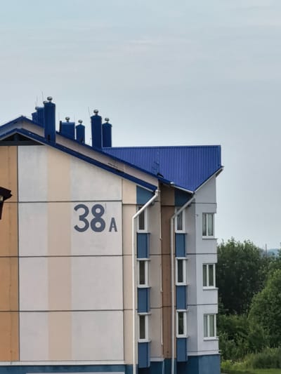 A building with a blue metal roof is visible against a pale blue sky. The building has multiple stories, with a section on the left featuring vertical bands of yellow and white. A large, dark blue number "38" is painted on this section, with a smaller "A" next to it. To the right, another section of the building is visible, painted in white and light blue vertical stripes, with several windows. Several blue chimneys with silver caps are on the roofline. In the background, lush green trees and foliage are visible. The overall scene suggests a residential building in a suburban or semi-urban environment.