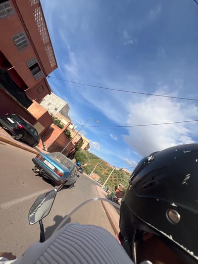 The image, taken from the perspective of a person riding a motorcycle, shows a street scene in Ouaouizert, Morocco. A black helmet is prominent in the foreground, partially obscuring the view. The street curves slightly to the right. Parked along the side of the road are a blue, older model car with a visible license plate number "32543 T 61", and a black hatchback car. Buildings in the background are predominantly reddish-brown and light gray in color. Utility lines are visible against a blue sky with scattered clouds. A hillside with olive trees is also visible in the background. The road surface appears to be asphalt. The motorcycle's rearview mirror is partially visible on the left.