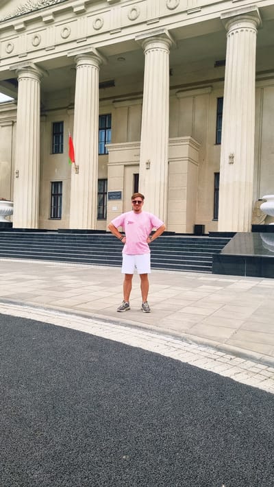 A full-shot outdoor, eye-level photograph captures a young man standing in front of a grand, neoclassical building. The man, wearing a pink t-shirt and white shorts, stands with his hands on his hips, facing the camera. He has sunglasses perched on his nose and is looking directly at the viewer. Behind him, the building features large, fluted columns supporting an ornate entablature. A Belarusian flag is visible hanging from a pole on the left side of the building. The ground in front of the building is paved with large square tiles, and a section of dark asphalt road is visible in the foreground. The scene is bright and appears to be daytime. This building is likely the Grodno State Agrarian University in Hrodna, Belarus.
