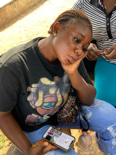A person rests their chin on their hand while looking at a smartphone. The phone displays a social media profile with several videos or photos. The person wears a black T-shirt with a large graphic on it and sits next to someone in a black and white striped shirt. Light blue jeans are also visible. A black handbag sits on the person's lap. The setting appears to be outdoors in daylight.